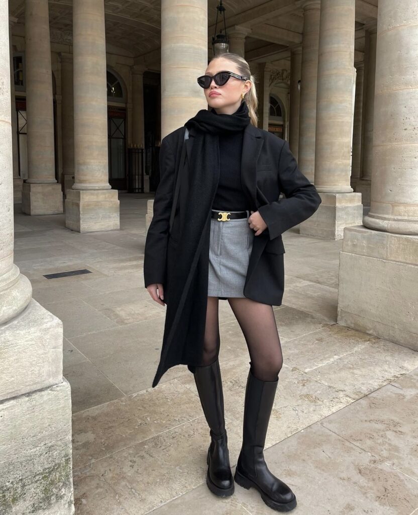 A sleek, professional winter outfit featuring a black blazer, grey mini skirt, and knee-high boots, complemented by a black scarf and sunglasses for a chic look.