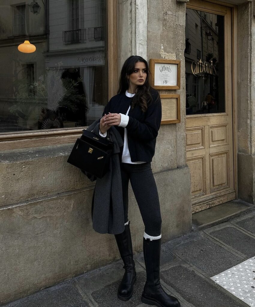 A cozy, layered winter look with black leggings, knee-high boots, a white sweater, and a black jacket.