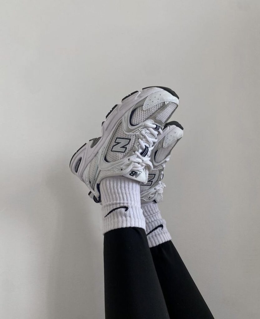 A pair of New Balance 530 sneakers worn with black leggings and white socks, legs crossed and held up against a plain white wall.