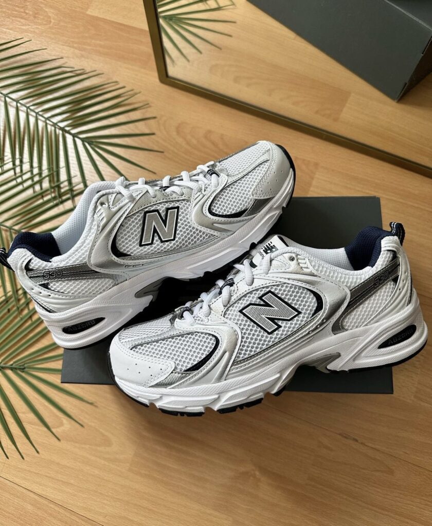 A close-up of a pair of New Balance 530 sneakers, showcasing the white and silver mesh design with the iconic 'N' logo. The sneakers are displayed on a wooden floor with a palm leaf accent in the background.