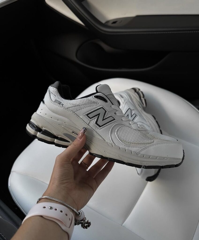 A hand holding a pair of New Balance sneakers in a car, highlighting the sleek and modern design of the shoes. The white and black colorway makes them versatile for everyday wear.