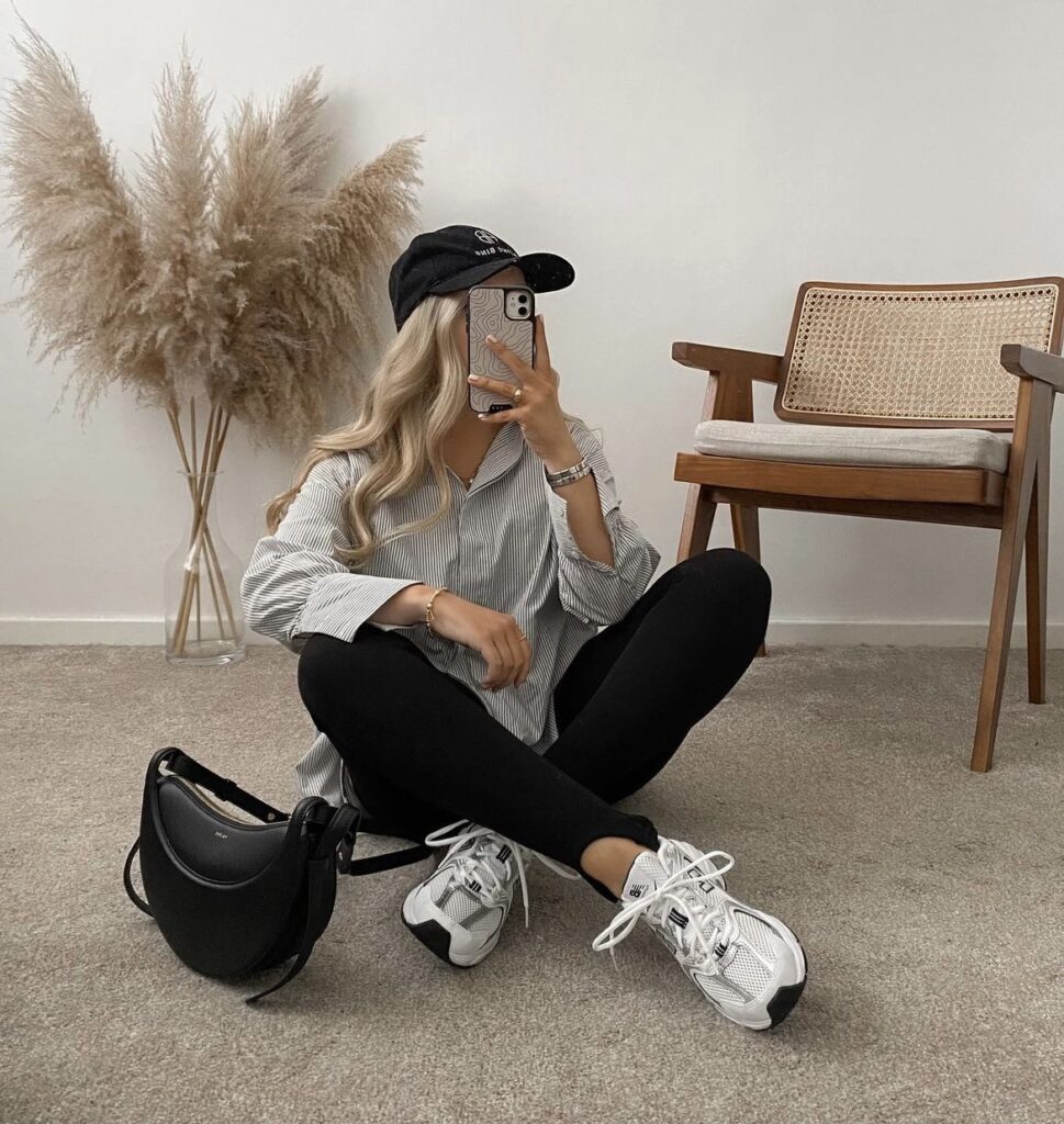 A person seated on the floor, dressed in a relaxed outfit consisting of a striped shirt, black leggings, and a black cap. White sneakers and a black handbag complete the comfortable and laid-back look.