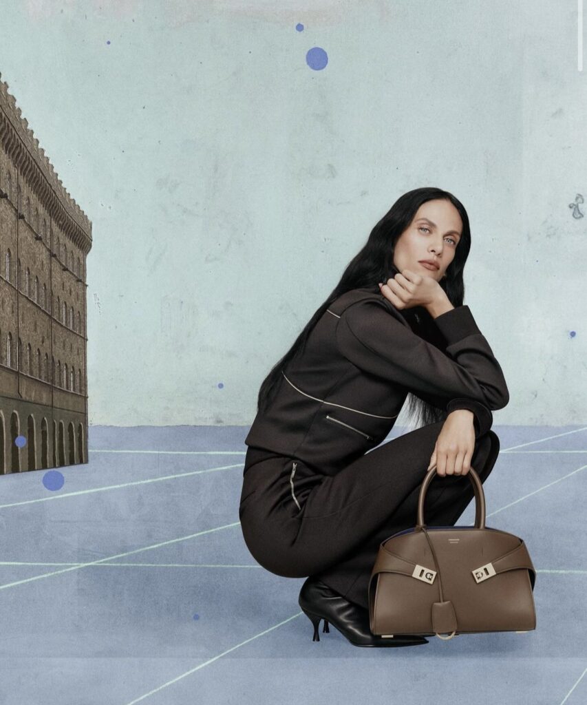 A model dressed in black, holding a brown Ferragamo handbag. The model is crouched in front of an architectural building.