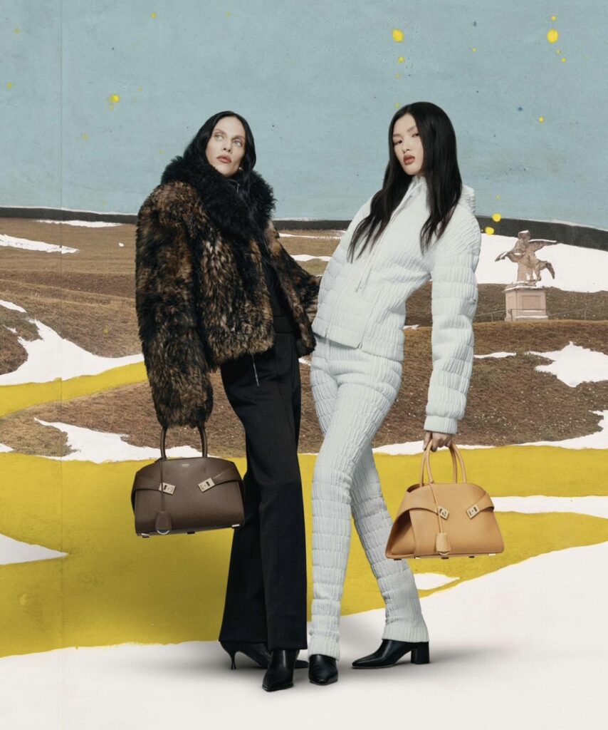 Two models pose in a wintery outdoor setting. One model wears a fur coat and black pants, holding a brown Ferragamo handbag, while the other wears a white quilted outfit, holding a tan Ferragamo handbag. The background features snow-covered fields and a statue in the distance.