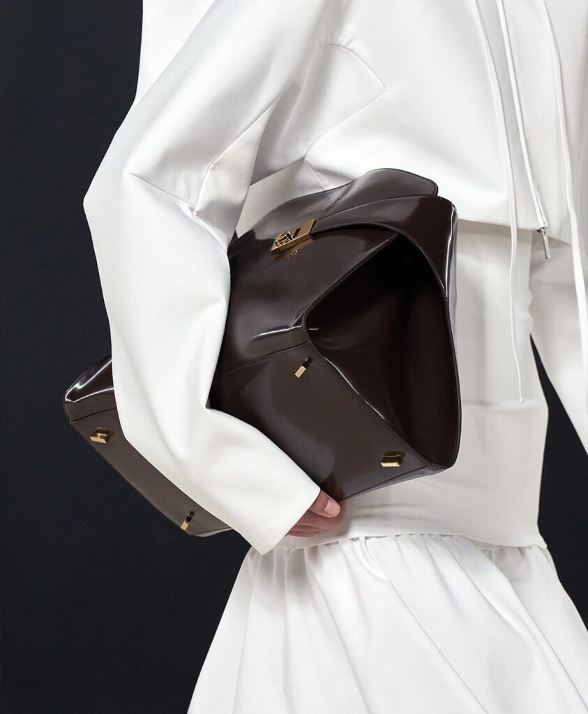 A close-up shot of a model holding a glossy dark brown Ferragamo handbag against a black backdrop. The model is wearing a white outfit.